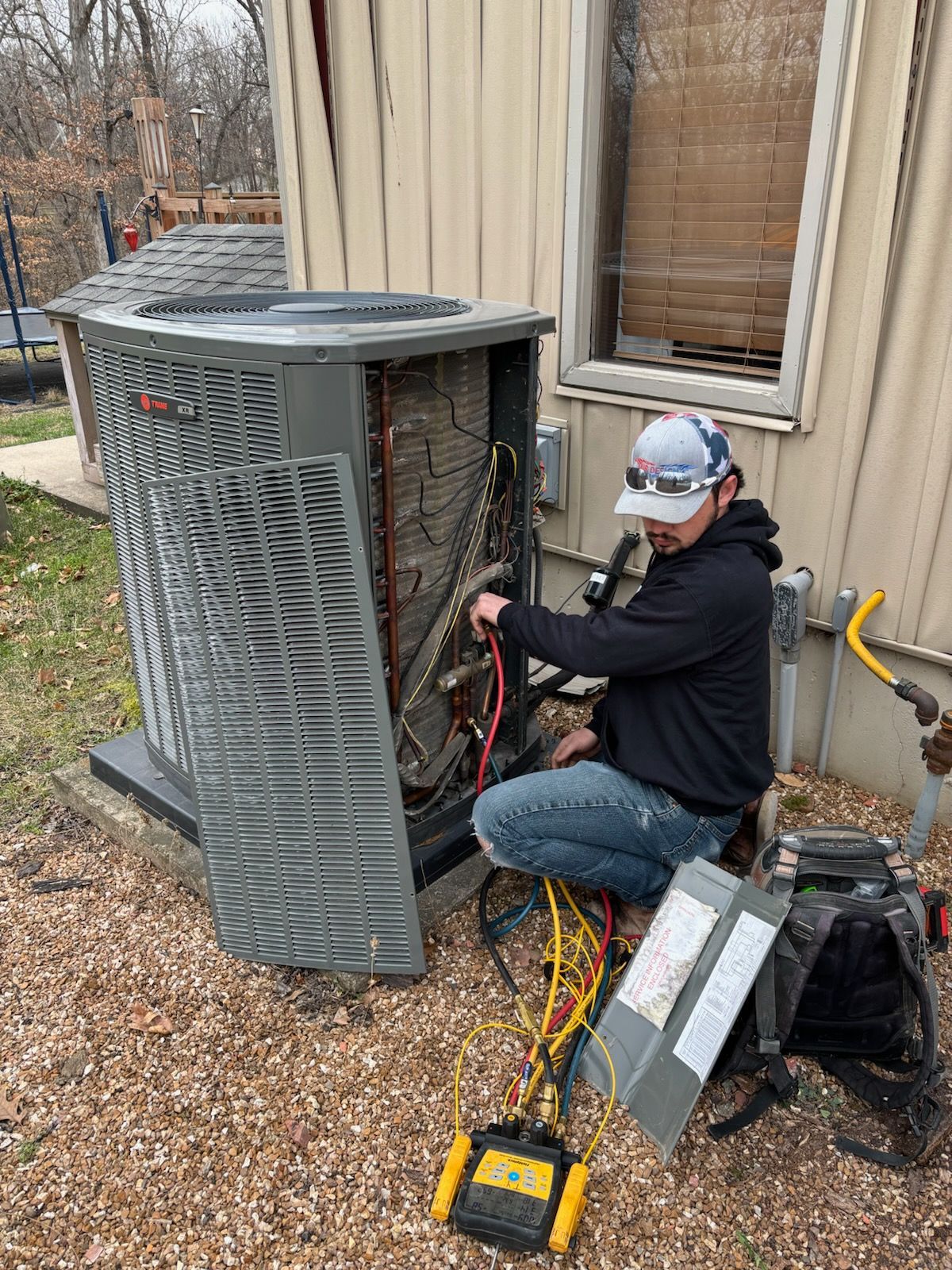 HVAC technician repairing an air conditioner unit outside a house, using tools and wires.