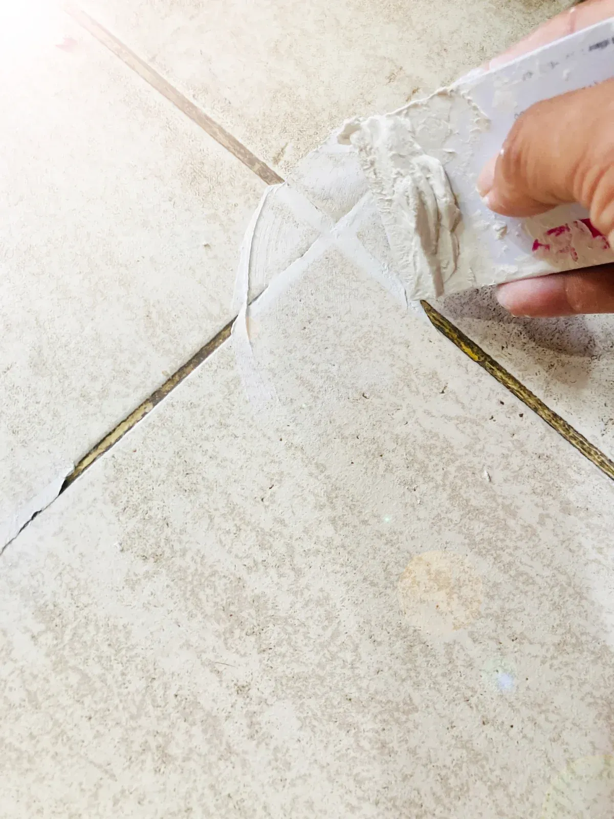 Hand applying grout with a trowel to fill tile seams on a light-colored floor.