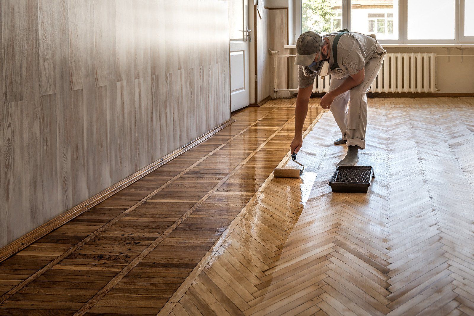 Man applying varnish to herringbone hardwood floor, wearing mask, in a room with light walls.
