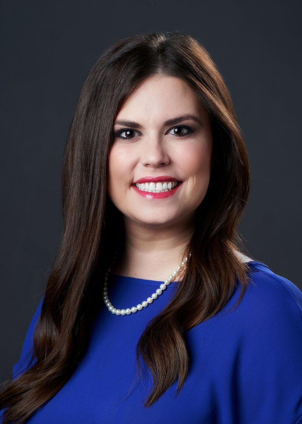 A woman wearing a blue shirt and a pearl necklace is smiling for the camera.