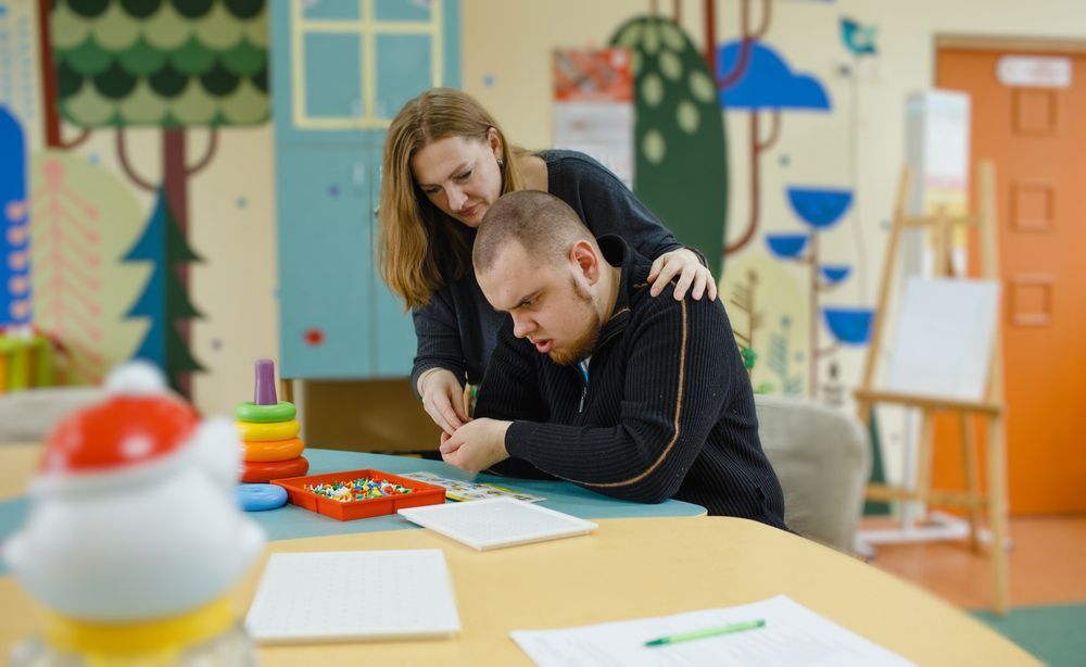 A woman is standing next to a man sitting at a table.