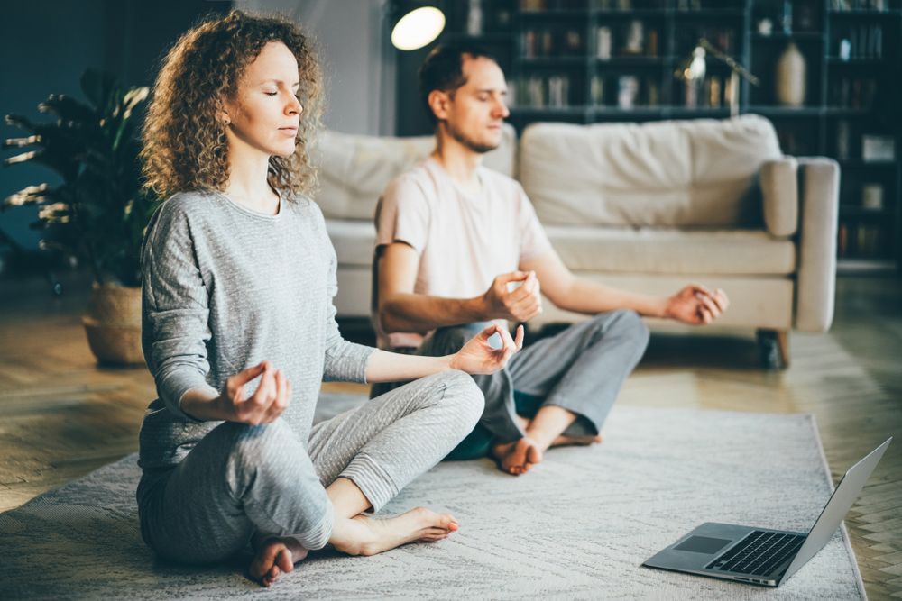 A man and a woman are sitting on the floor meditating in front of a laptop.