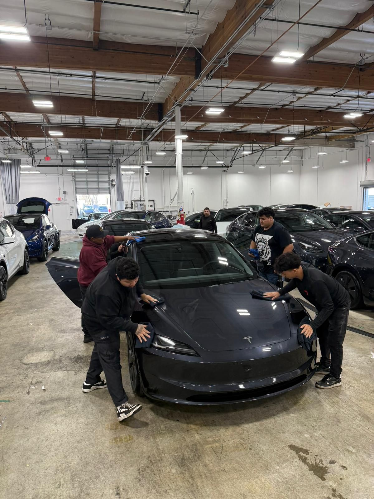 People cleaning a dark gray Tesla inside a garage.