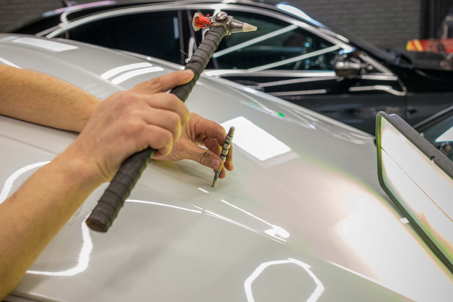 Hands using a hammer and tool to repair a dent in a silver car.