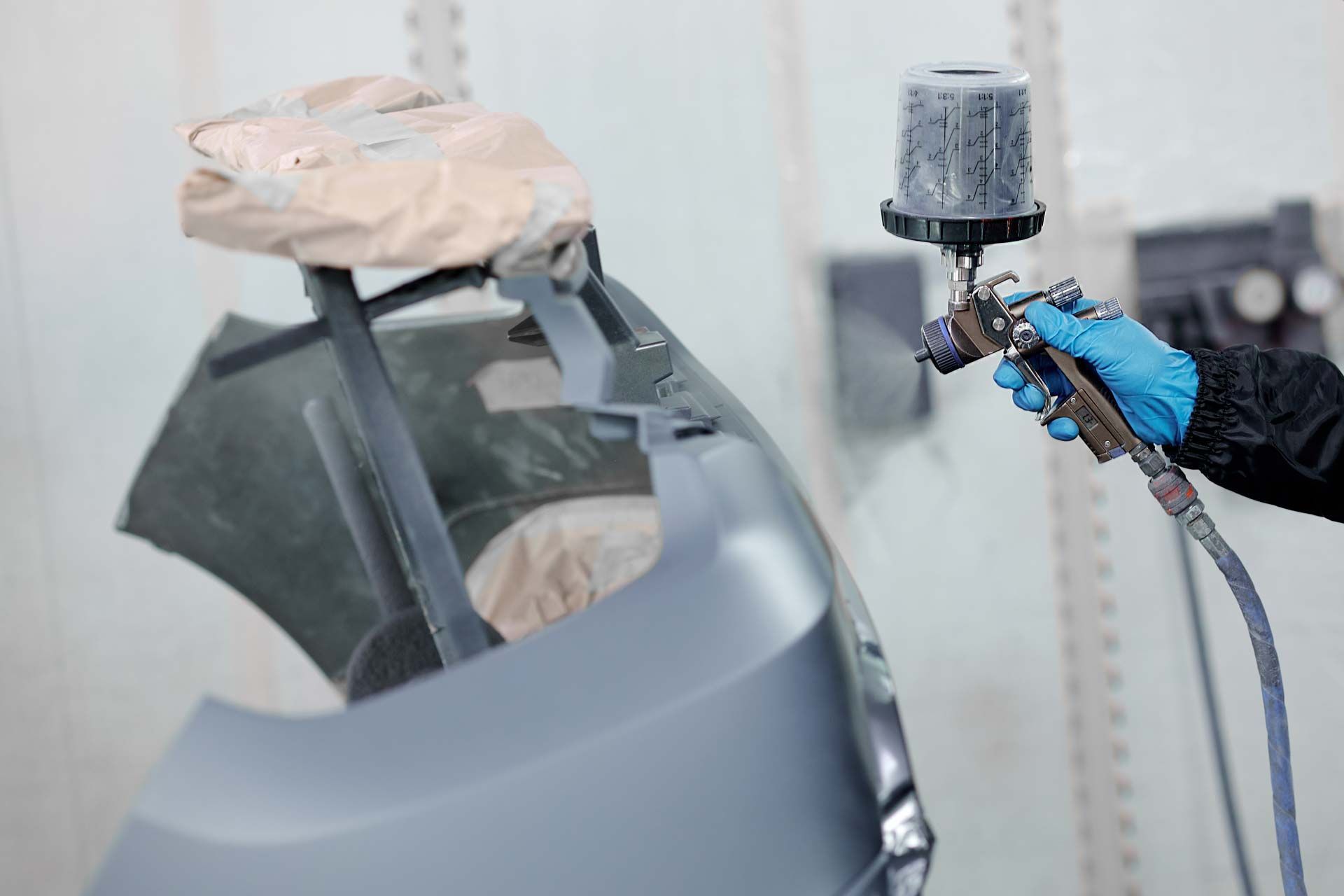 A person in blue gloves sprays paint on a car bumper in a workshop.
