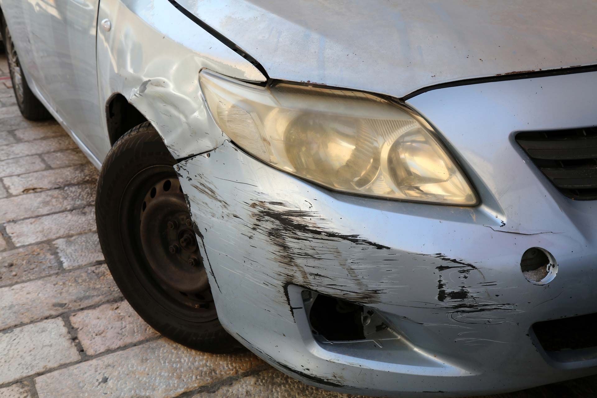 Damaged silver car bumper with scratches, a dent, and a clouded headlight.