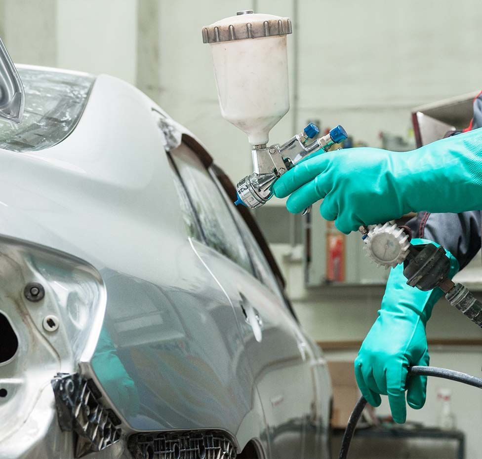 Person spraying silver paint on a car door in a garage, wearing teal gloves.