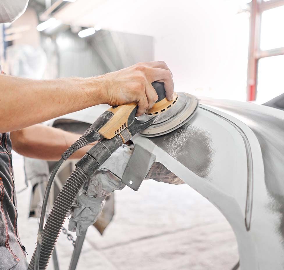 Person sanding a car bumper in a workshop, using a power sander.