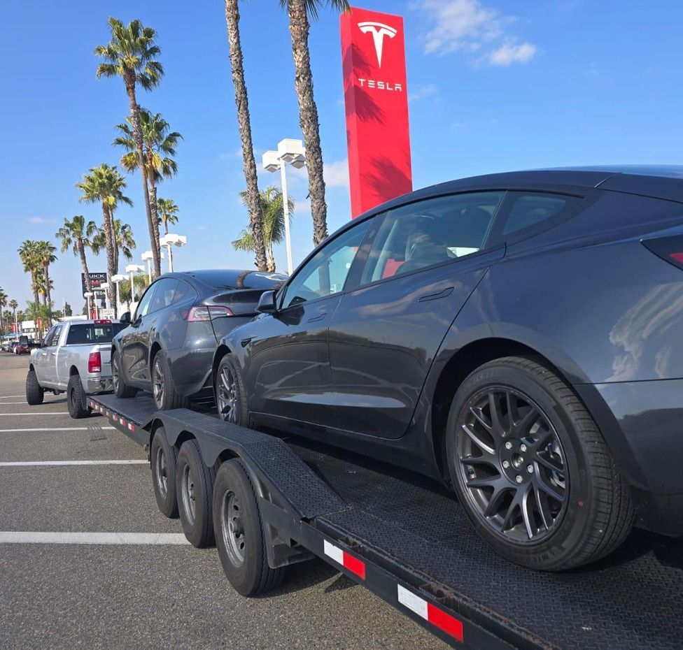 Two Tesla cars on a car hauler at a Tesla dealership, with a red Tesla sign and palm trees.