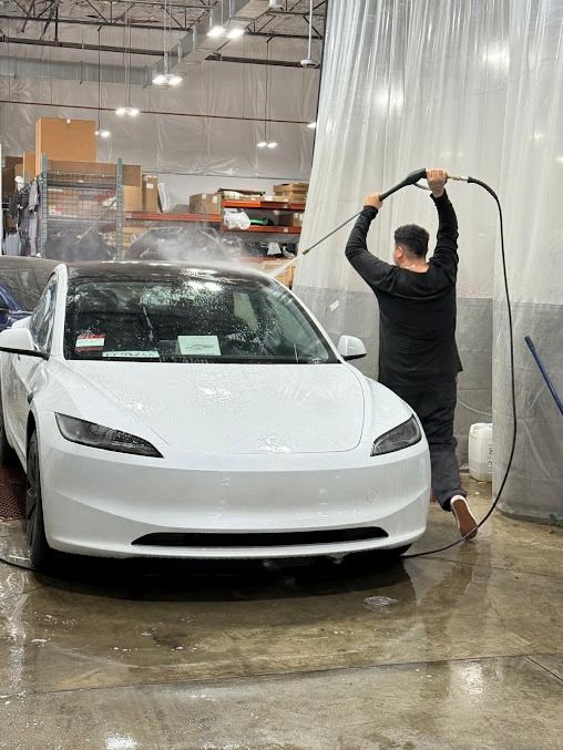 Man washing a white Tesla car with a hose indoors, water splashing on the vehicle and floor.