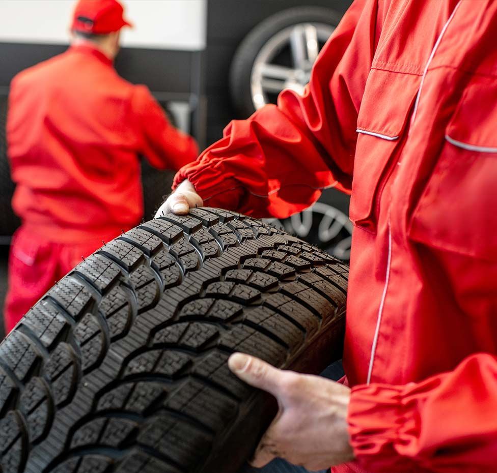 Mechanic holding a tire in a garage, another mechanic in the background, both wearing red jumpsuits.