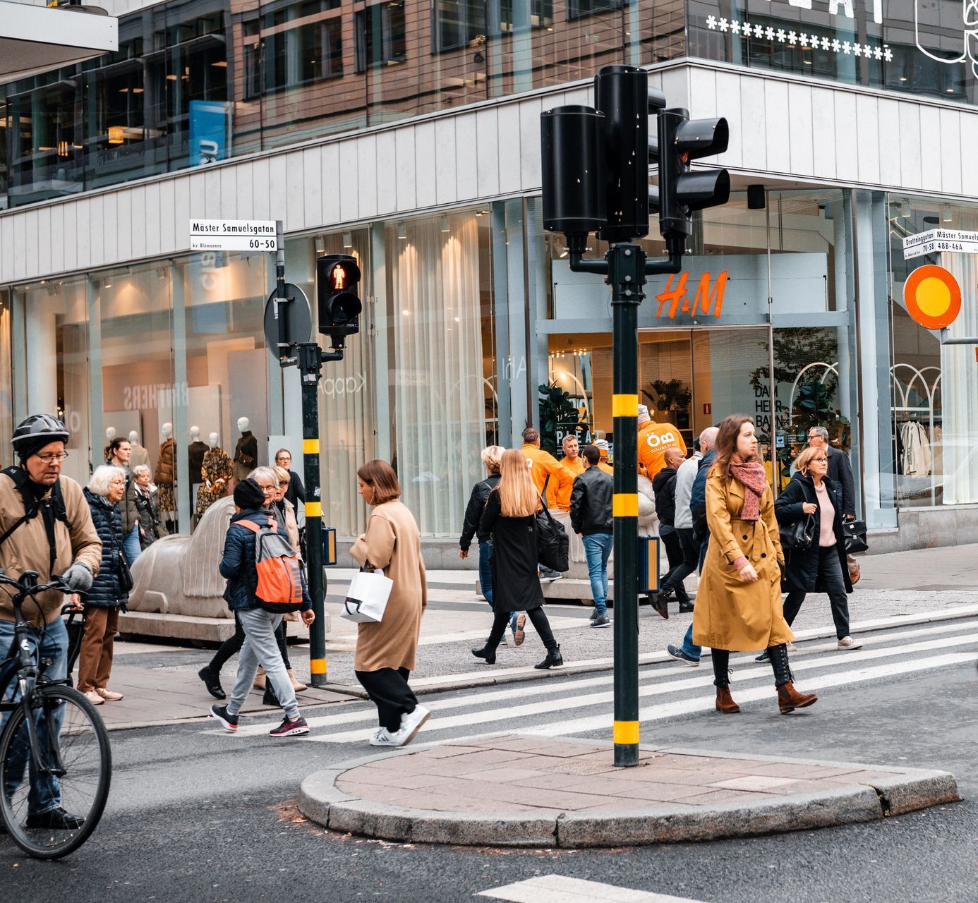 People cross a street at a crosswalk in front of an H&M store; traffic lights are visible.