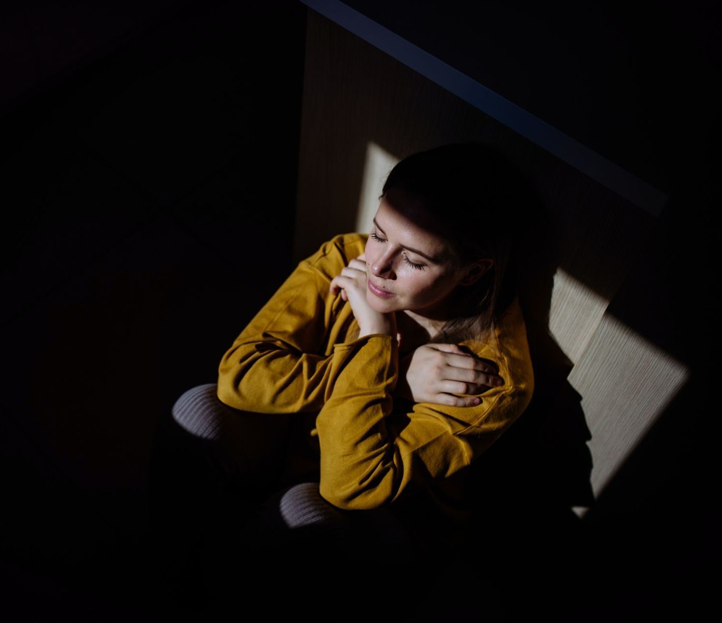 Woman in mustard shirt sits in a dark corner, lit by a patch of sunlight, looking contemplative.