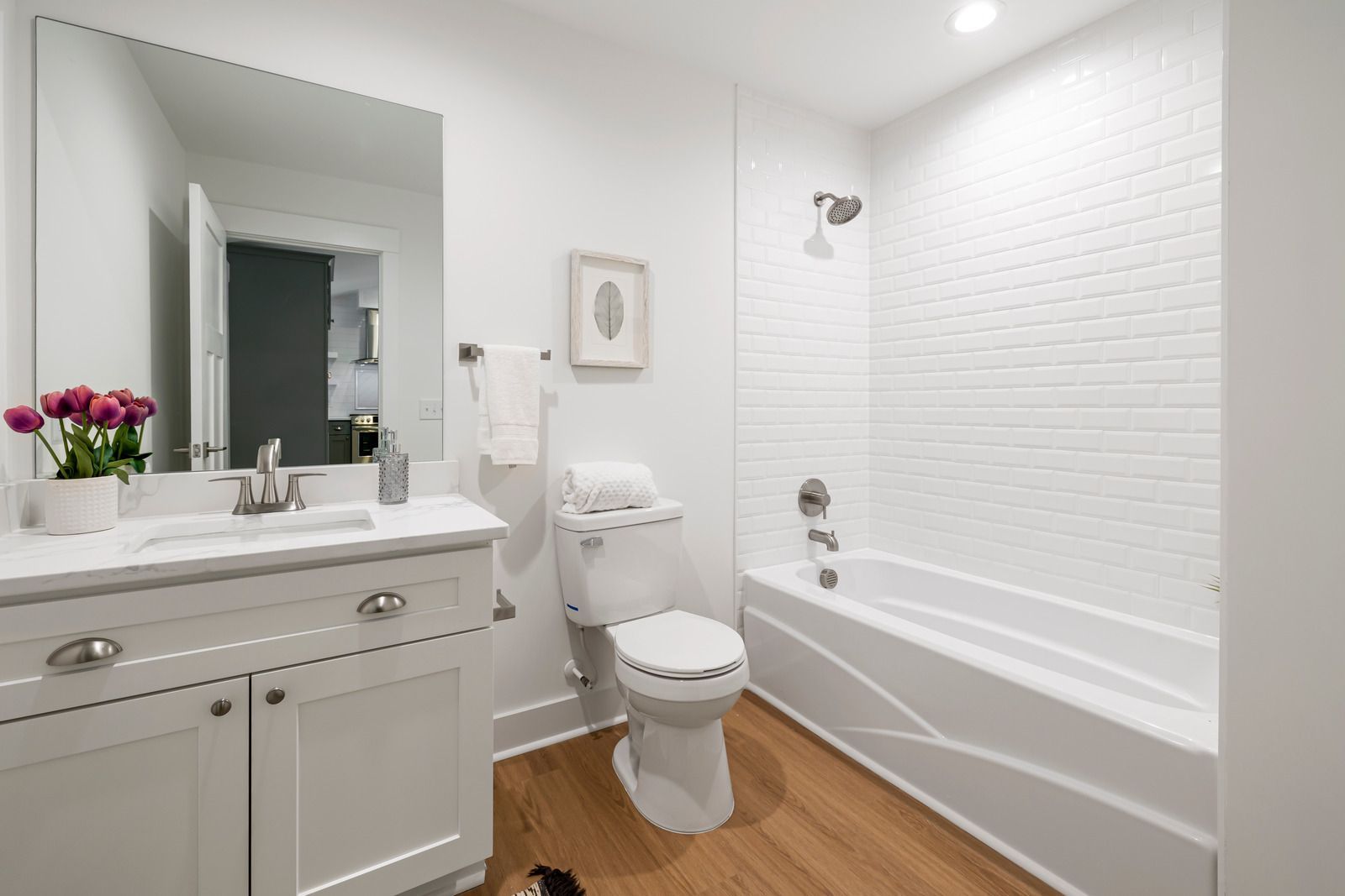 Bright white bathroom with vanity, mirror, toilet, and bathtub with white tile surround and wood floor