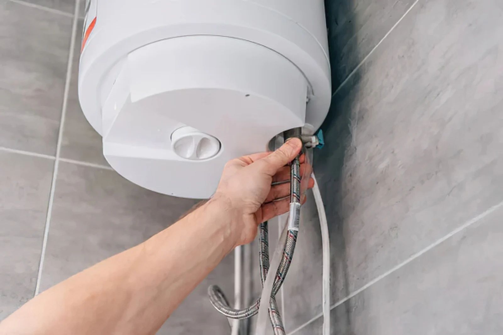 Hand connecting a chrome shower hose to a white water heater in a tiled bathroom