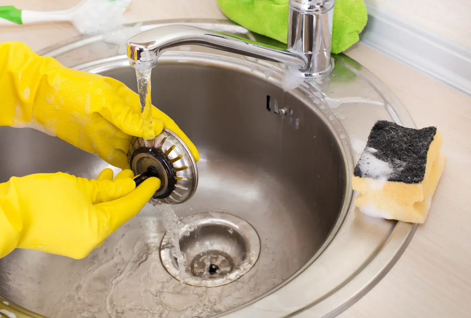 Hands in yellow gloves cleaning kitchen sink drain.