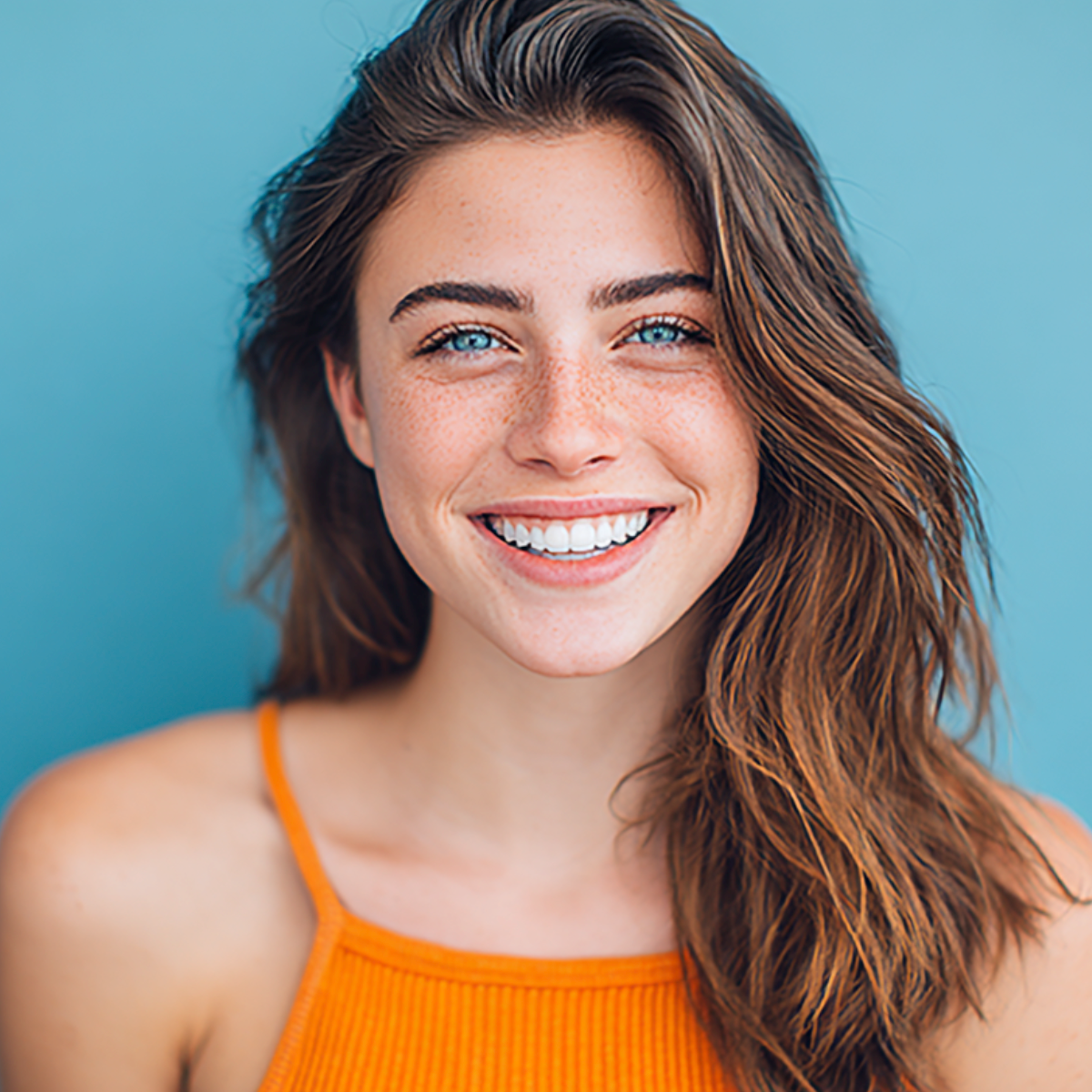 Woman with freckles smiles, blue eyes, brown hair, orange top against a blue backdrop.