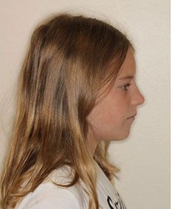 A young girl with long blonde hair is standing in front of a white wall.