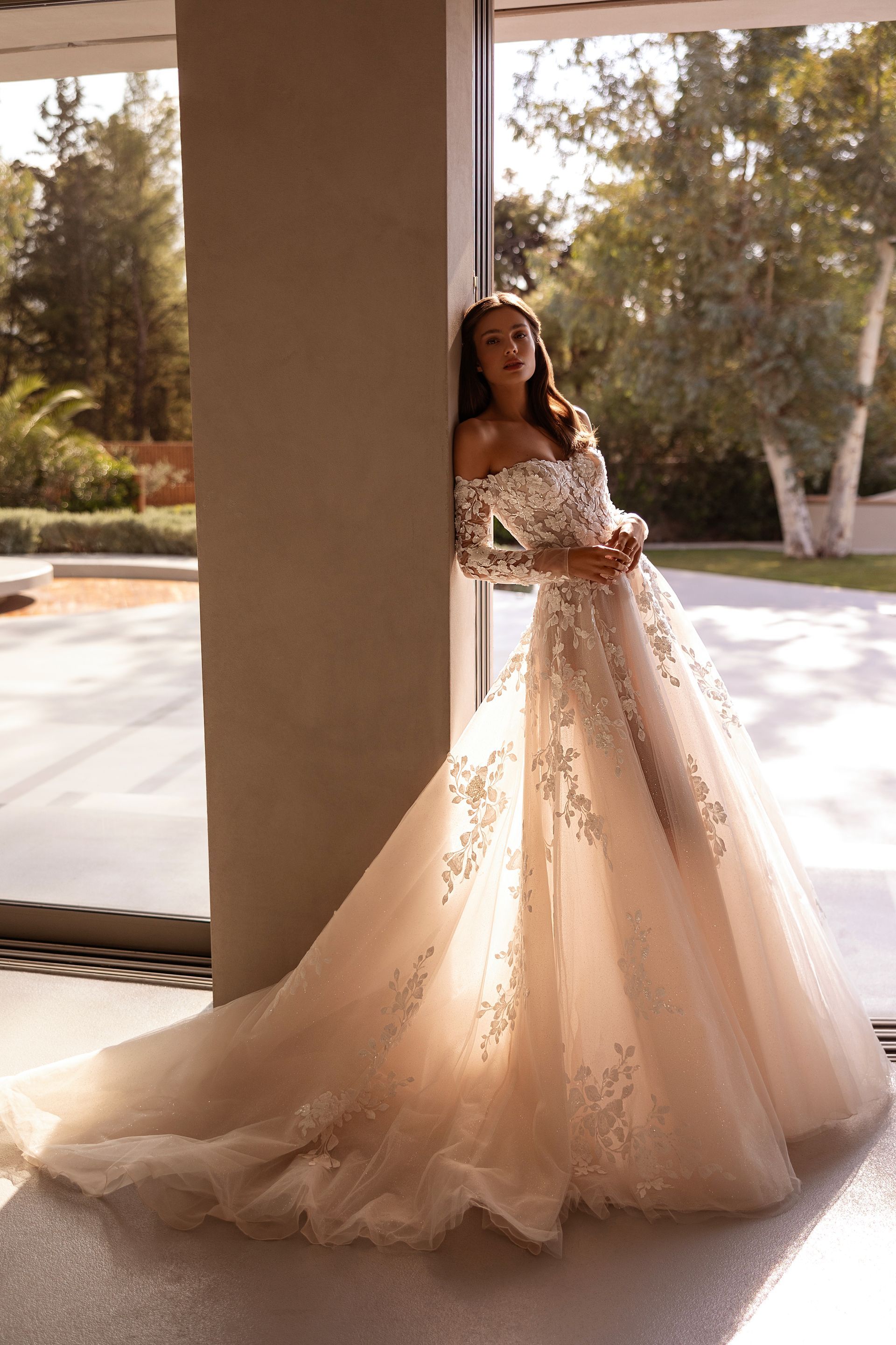 Donna con abito da sposa a spalle scoperte, appoggiata a un pilastro vicino a una finestra.