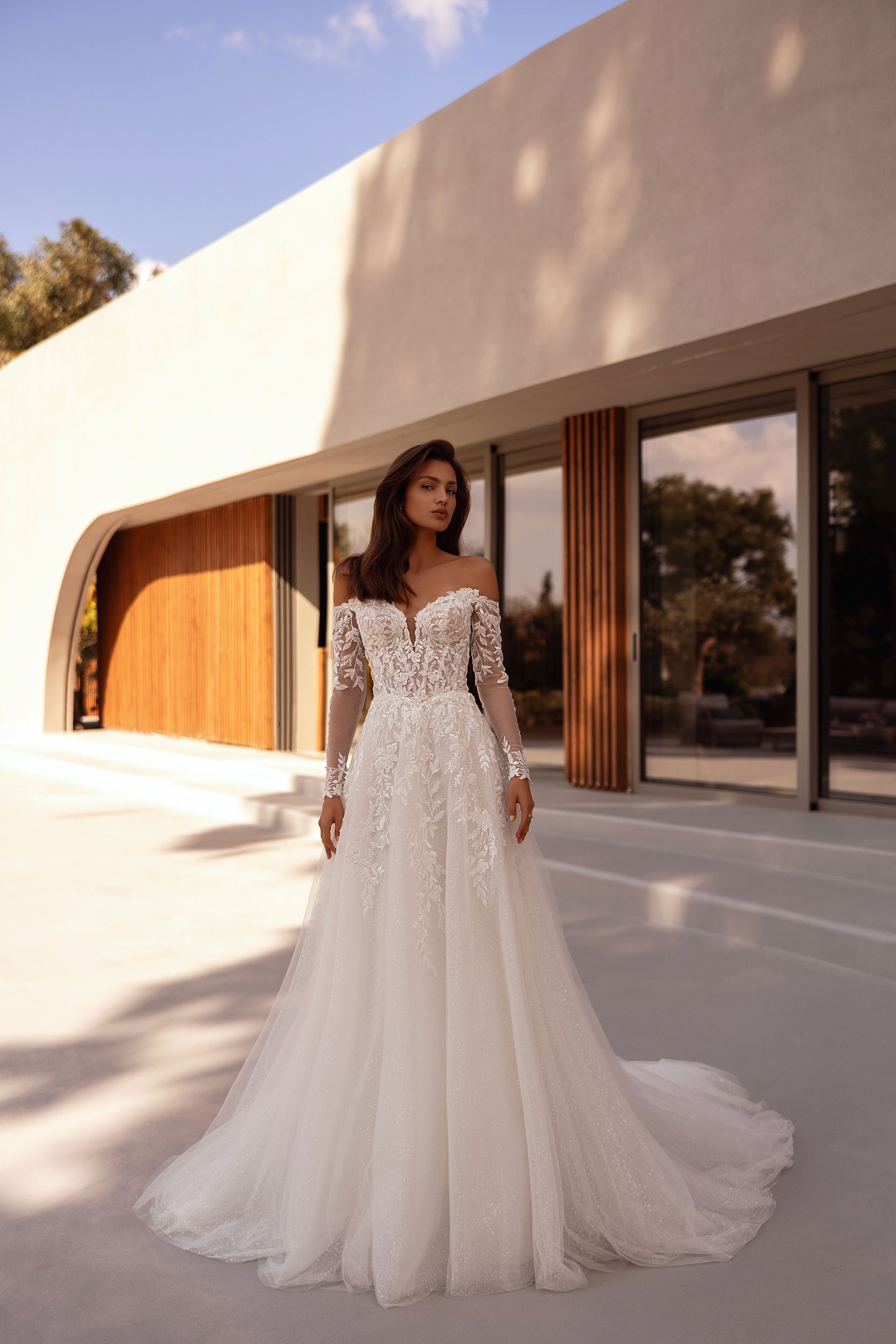 Donna con un abito da sposa in pizzo bianco con lungo strascico, in piedi fuori da un edificio moderno.