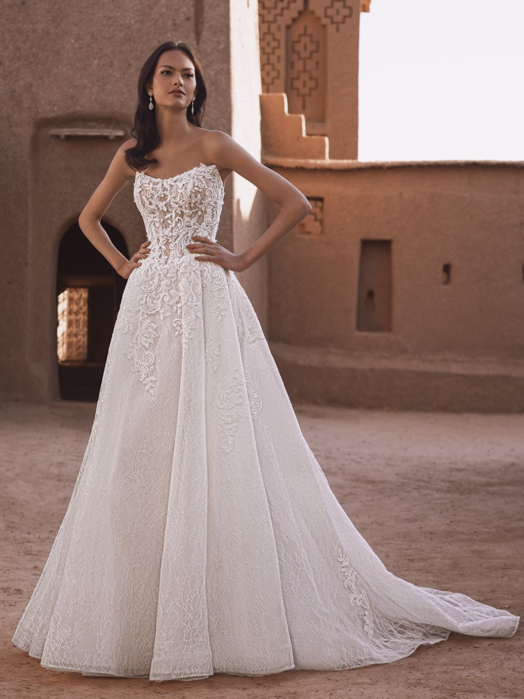 Donna con abito da sposa bianco senza spalline, mani sui fianchi, di fronte a un edificio strutturato.