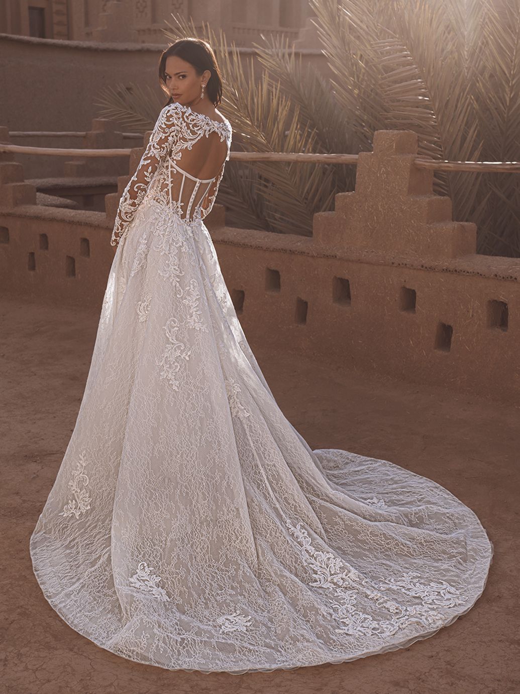 Donna in abito da sposa in pizzo bianco con lungo strascico, all'aperto vicino a una struttura in pietra arenaria.