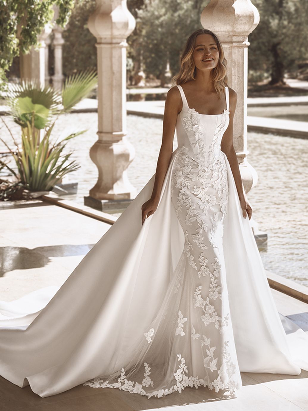 Donna sorridente all'aperto, in abito da sposa bianco con corpetto in pizzo e lungo strascico.