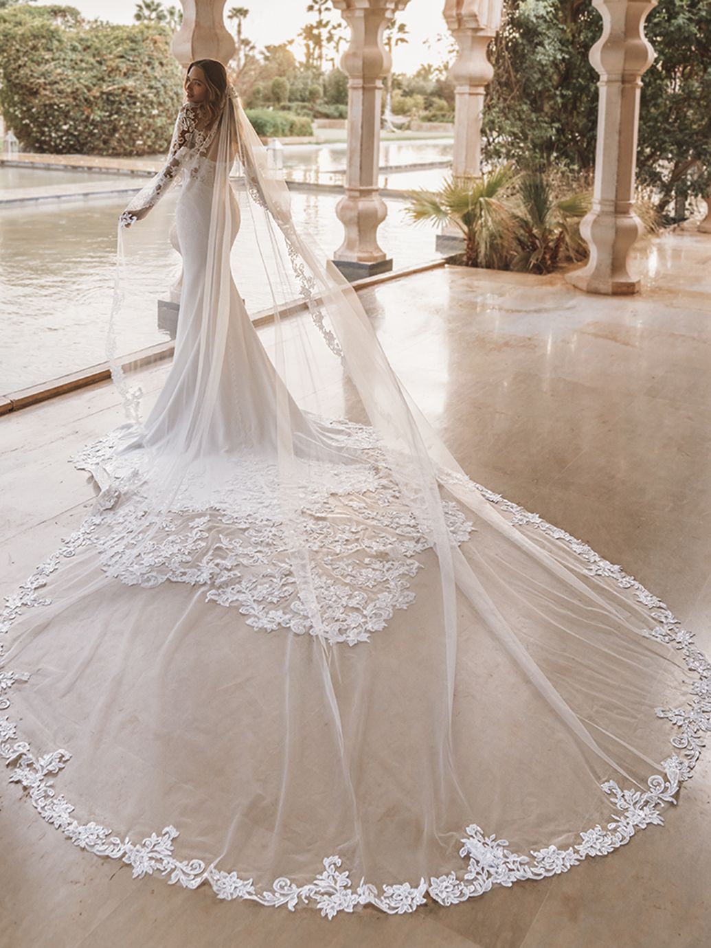 La sposa indossa un abito bianco con lungo strascico e velo ed è in piedi in un ambiente esterno coperto.