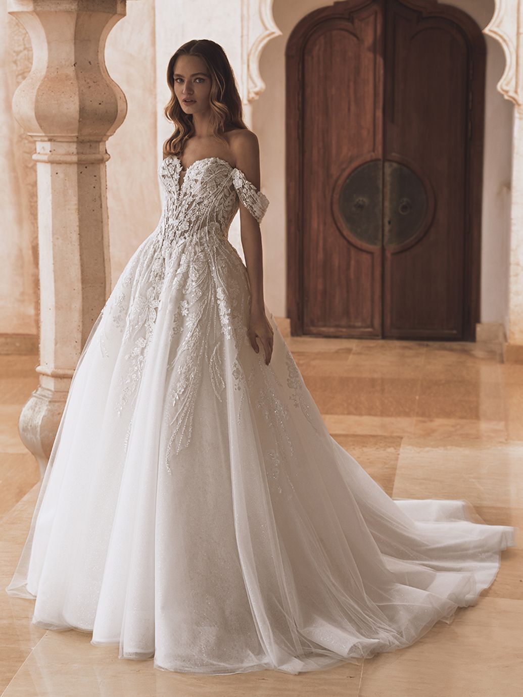Una donna con un abito da sposa bianco a spalle scoperte è in piedi in un ambiente sontuoso.