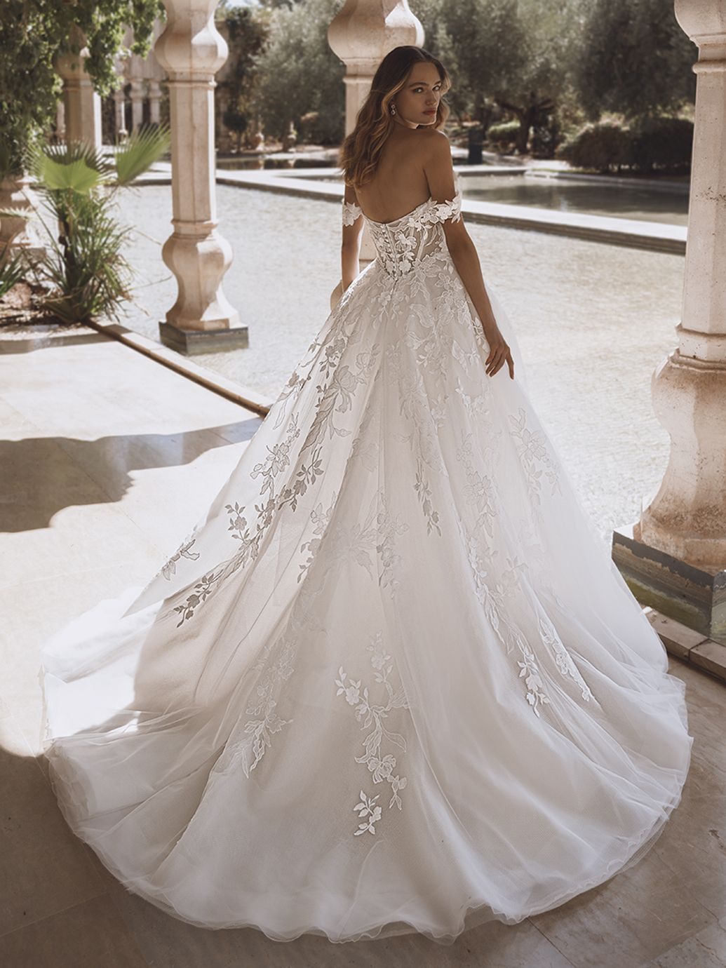 Donna in abito da sposa bianco con spalle scoperte, in piedi all'aperto vicino a una piscina riflettente e a dei pilastri.
