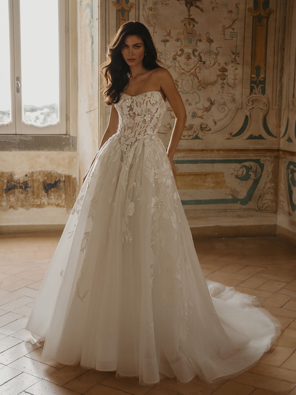 Donna in abito da sposa bianco senza spalline con dettagli in pizzo, in piedi in una stanza con pareti riccamente decorate.