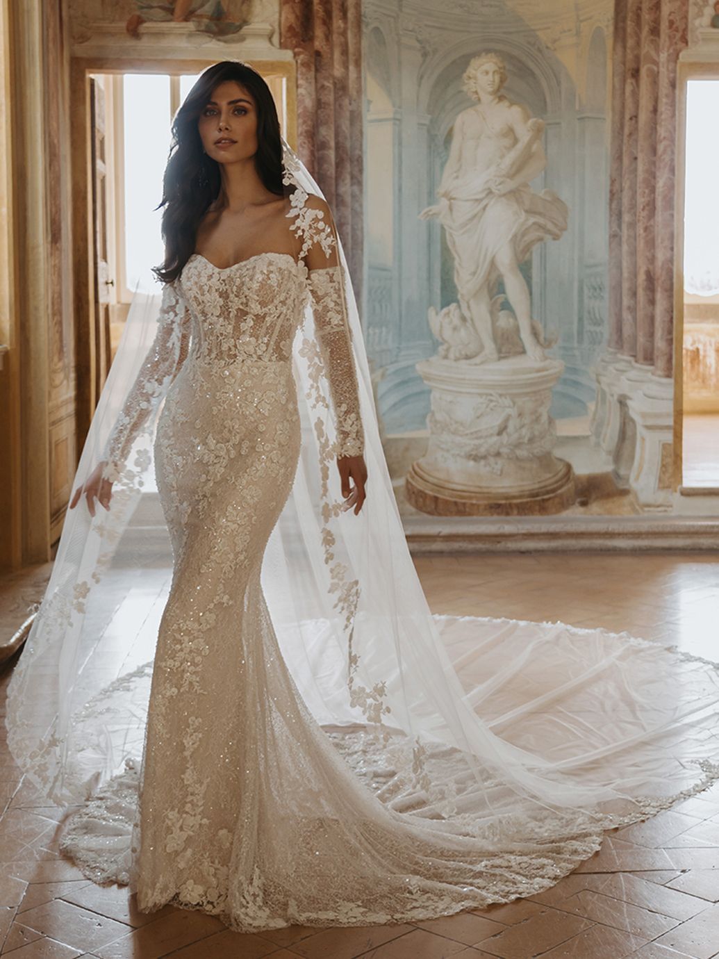 Donna in abito da sposa in pizzo bianco con lungo strascico, che regge il velo.