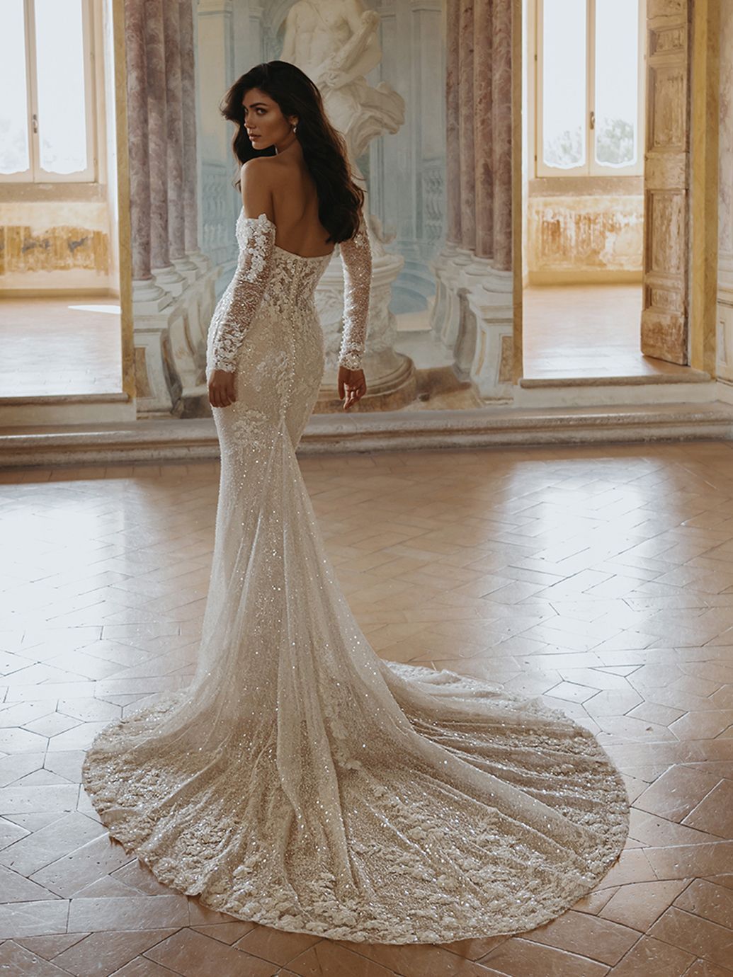 Donna in un abito da sposa scintillante, con spalle scoperte e lungo strascico, in piedi in una stanza ampia e luminosa.