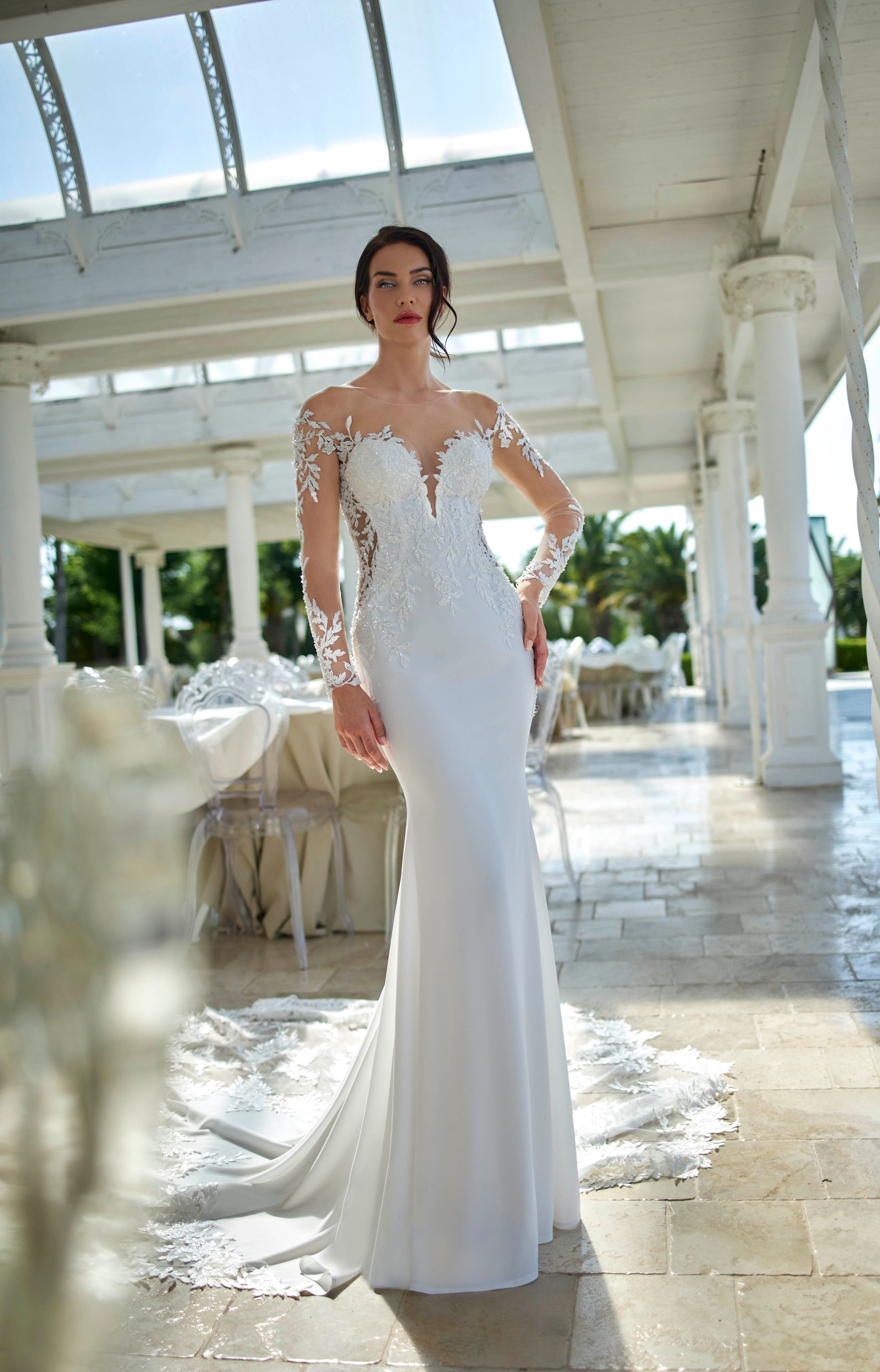 Donna in abito da sposa bianco a sirena con maniche in pizzo, in piedi in un ambiente soleggiato all'aperto.