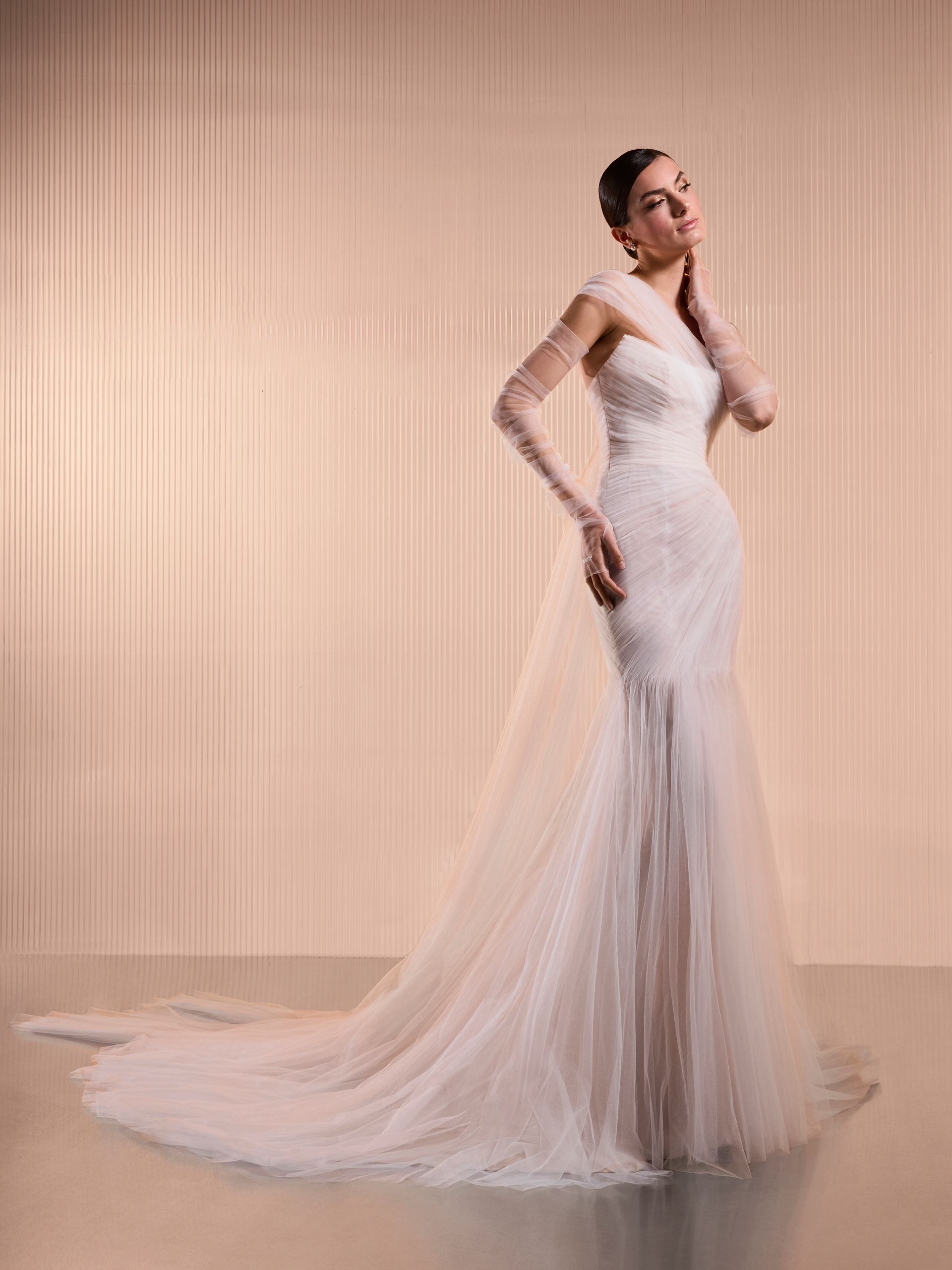 Donna in abito da sposa bianco a sirena con guanti trasparenti e lungo strascico, in piedi su uno sfondo strutturato.