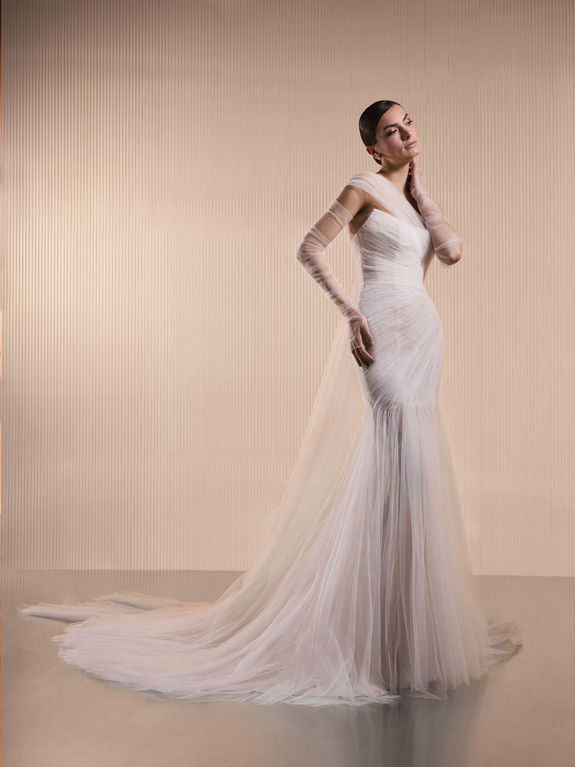 Donna in abito da sposa bianco con lungo strascico e guanti trasparenti, in posa contro un muro a coste.