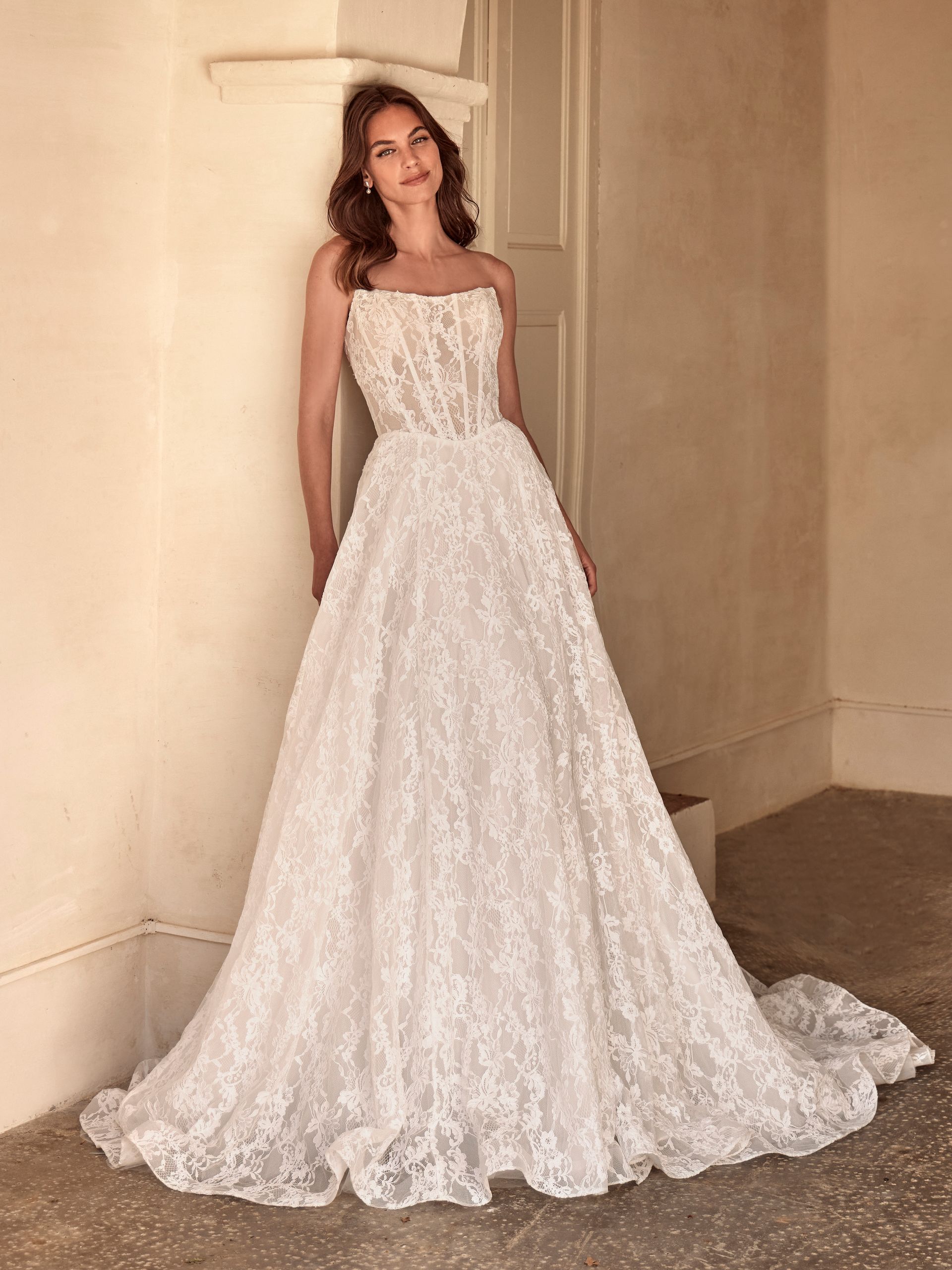 Donna in abito da sposa in pizzo bianco senza spalline appoggiata a un muro.