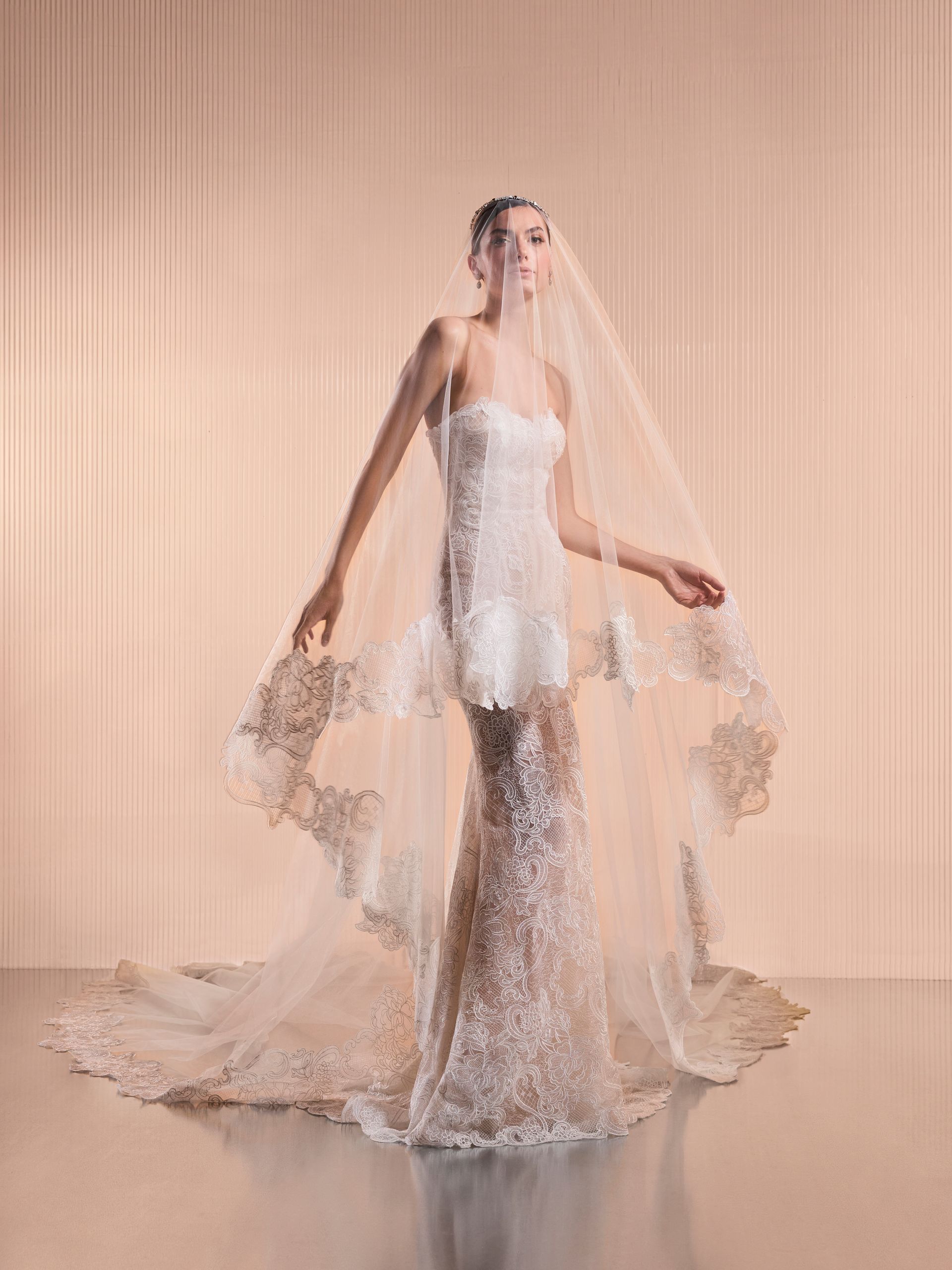 Sposa in abito senza spalline con decorazioni floreali e lungo velo di pizzo, in piedi davanti a uno sfondo strutturato.