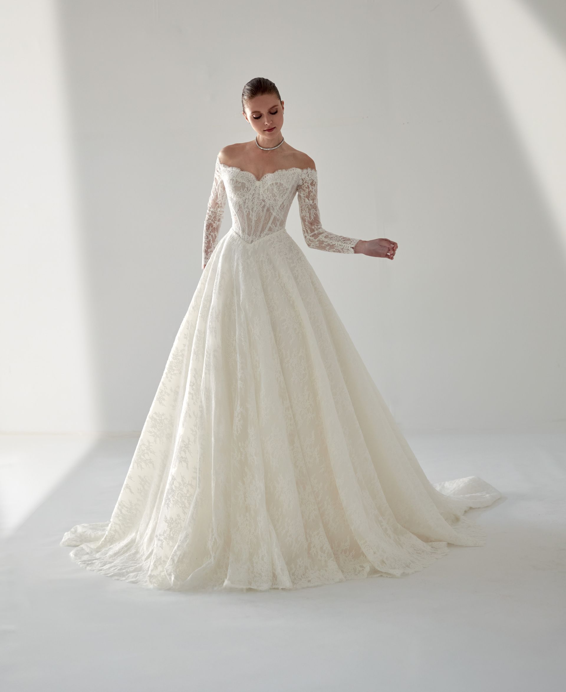 Donna in abito da sposa bianco con spalle scoperte e maniche lunghe. È in piedi in uno studio.