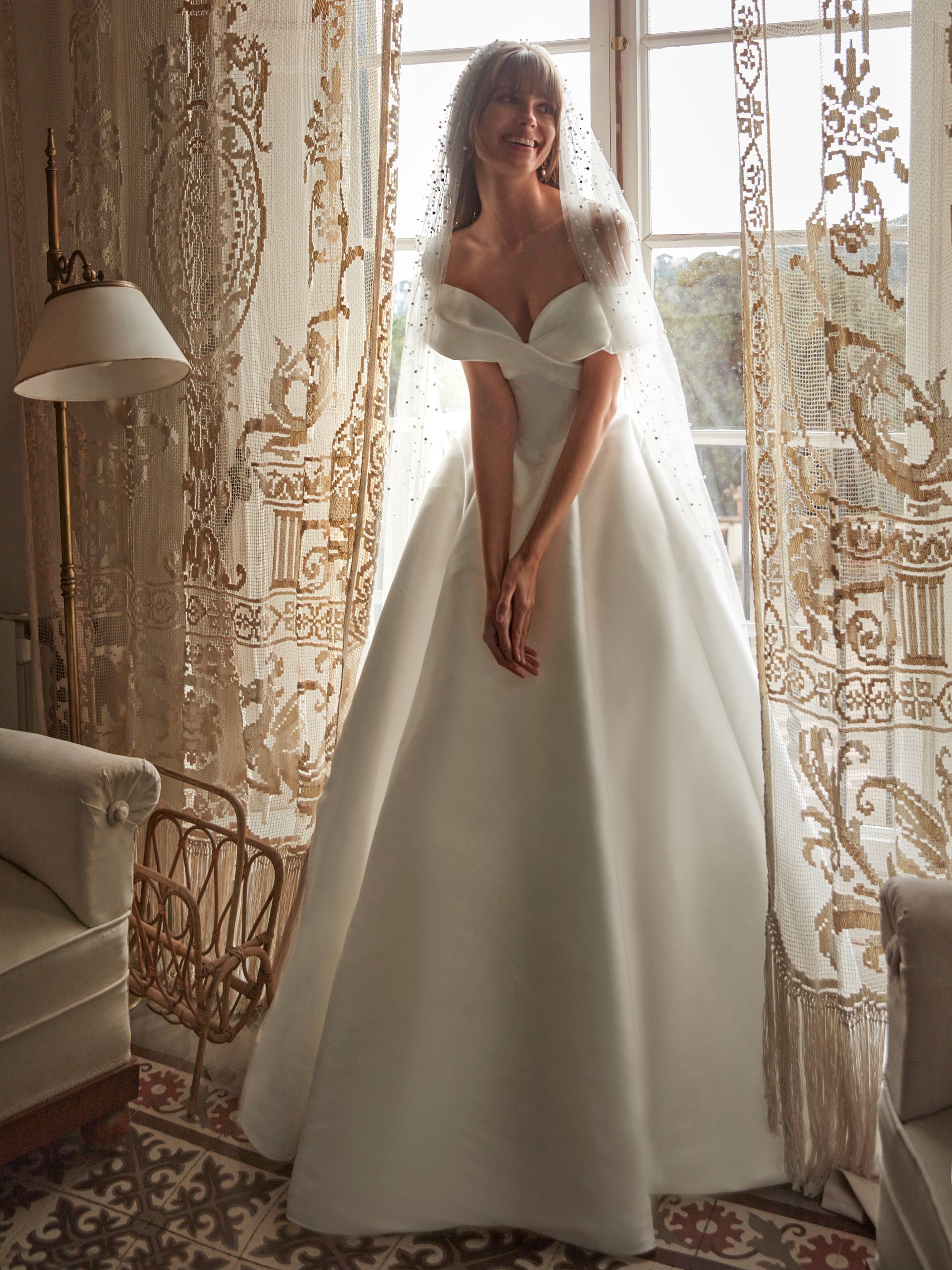 Donna in abito da sposa bianco con velo, sorridente vicino a una finestra con tende decorate.