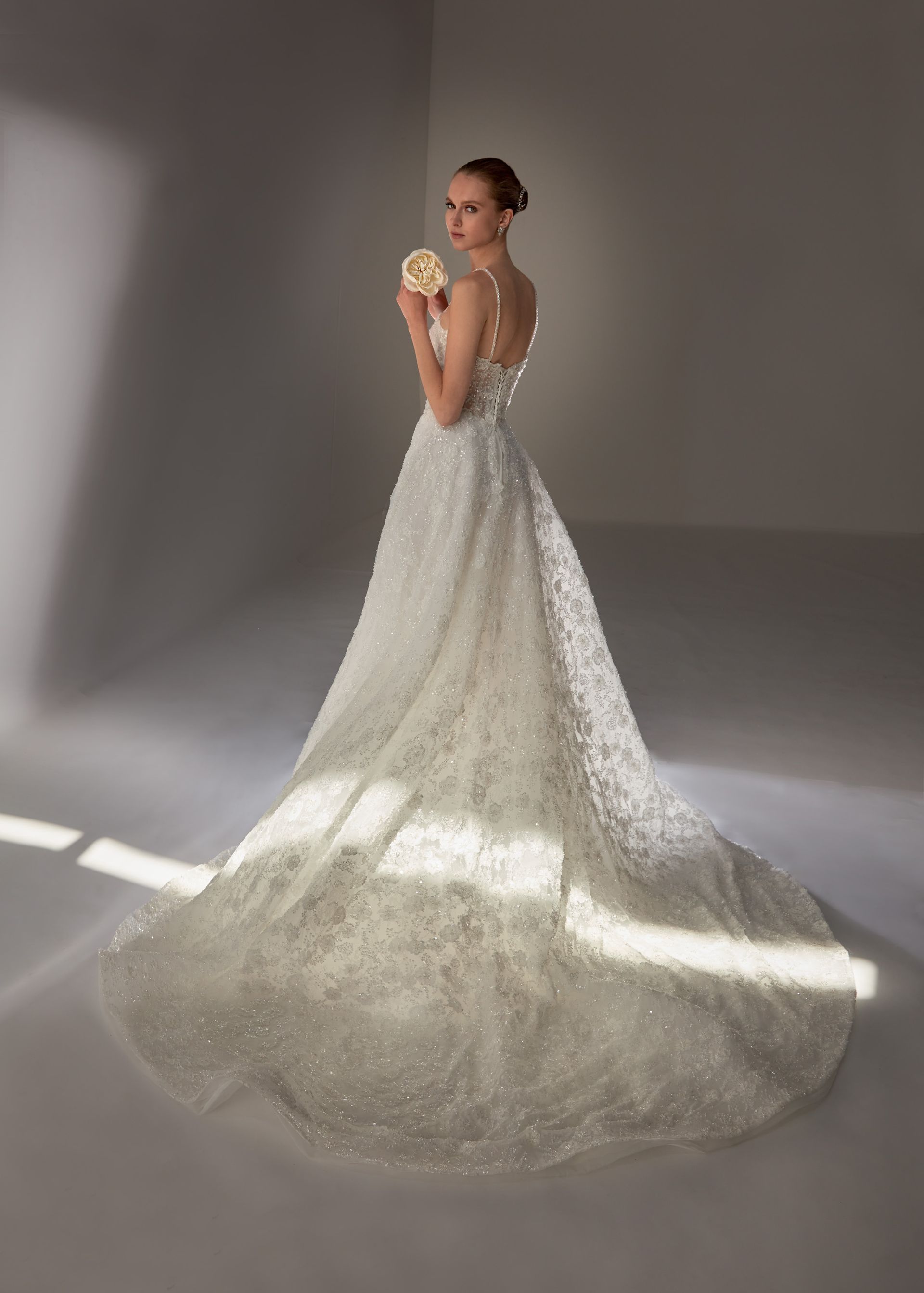 Donna in un abito da sposa bianco scintillante, con in mano un fiore, in piedi in uno studio illuminato dalla luce del sole.