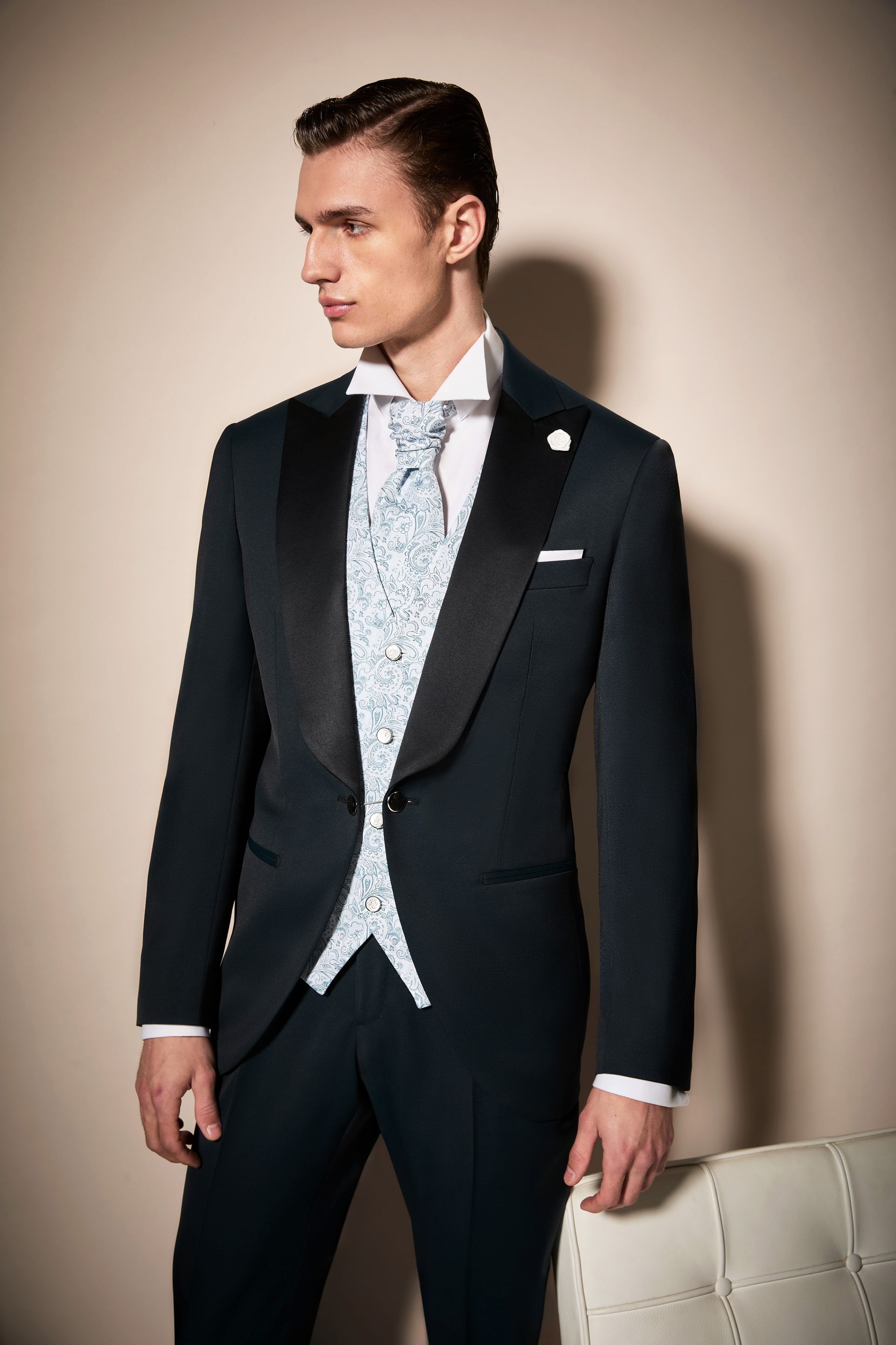 Uomo in un elegante abito blu scuro con gilet fantasia, camicia bianca e fiore all'occhiello, in posa in un interno.