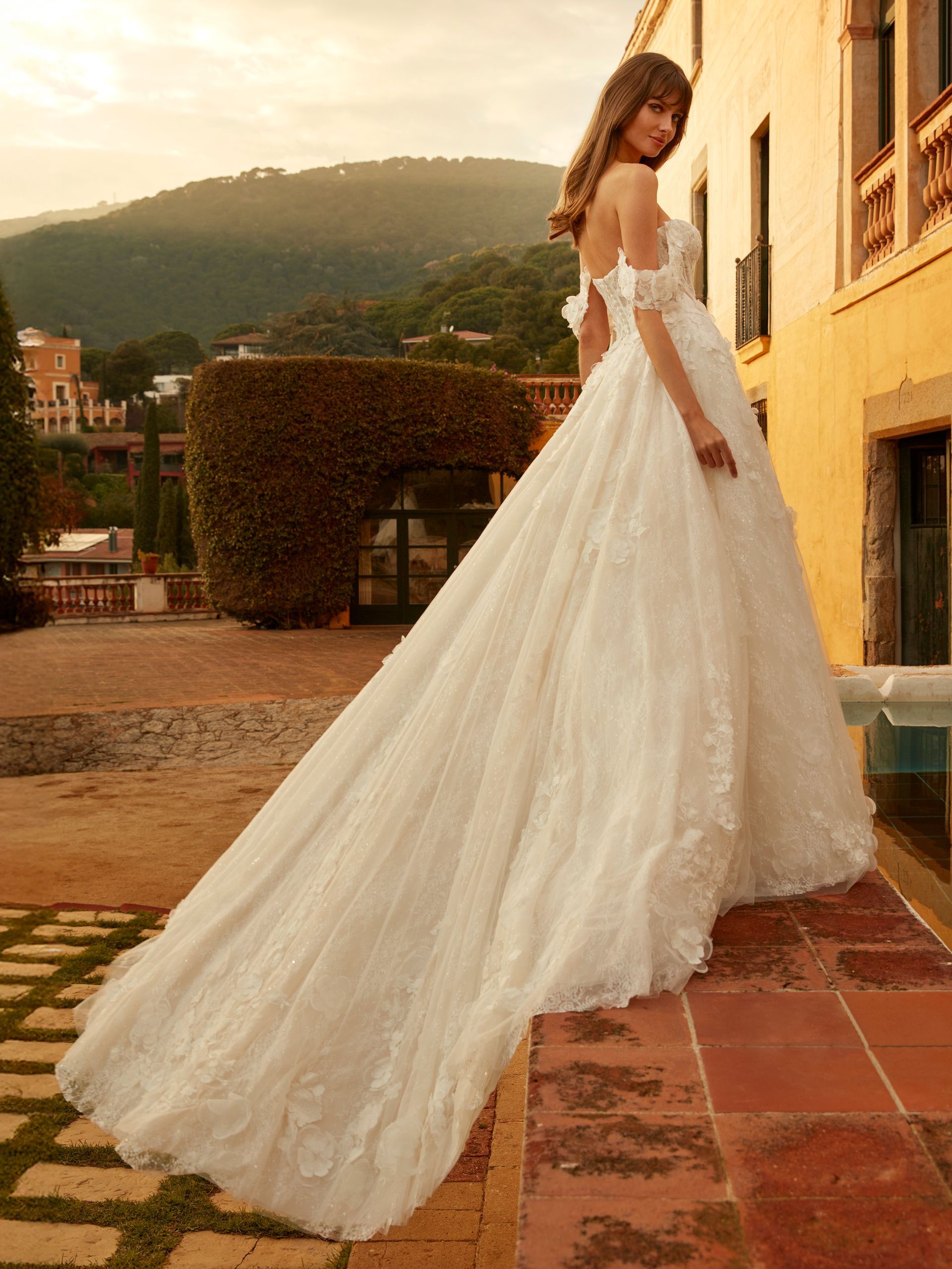 Una donna in abito da sposa bianco con lungo strascico è in piedi su un patio, con le montagne sullo sfondo.