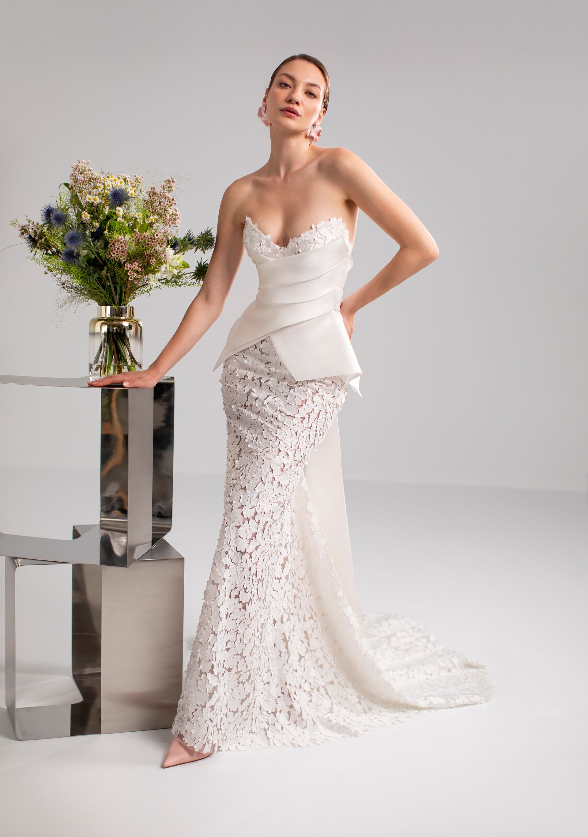 Donna in abito da sposa in pizzo bianco, in posa vicino a una composizione floreale su un tavolo a specchio.