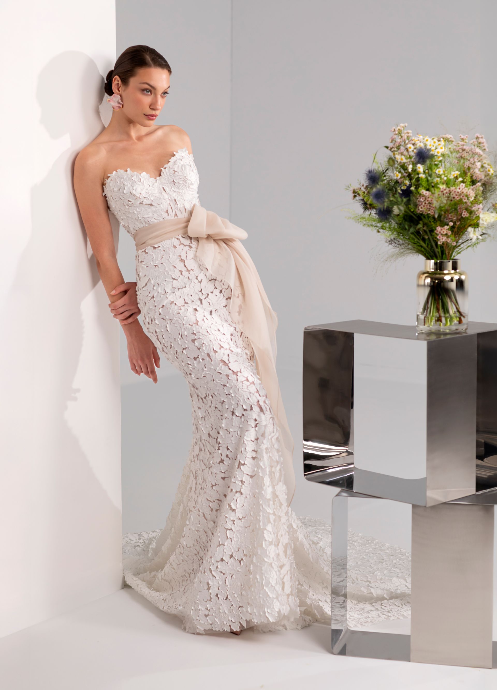 Una donna in un abito da sposa bianco senza spalline in pizzo è appoggiata a un angolo, in posa.