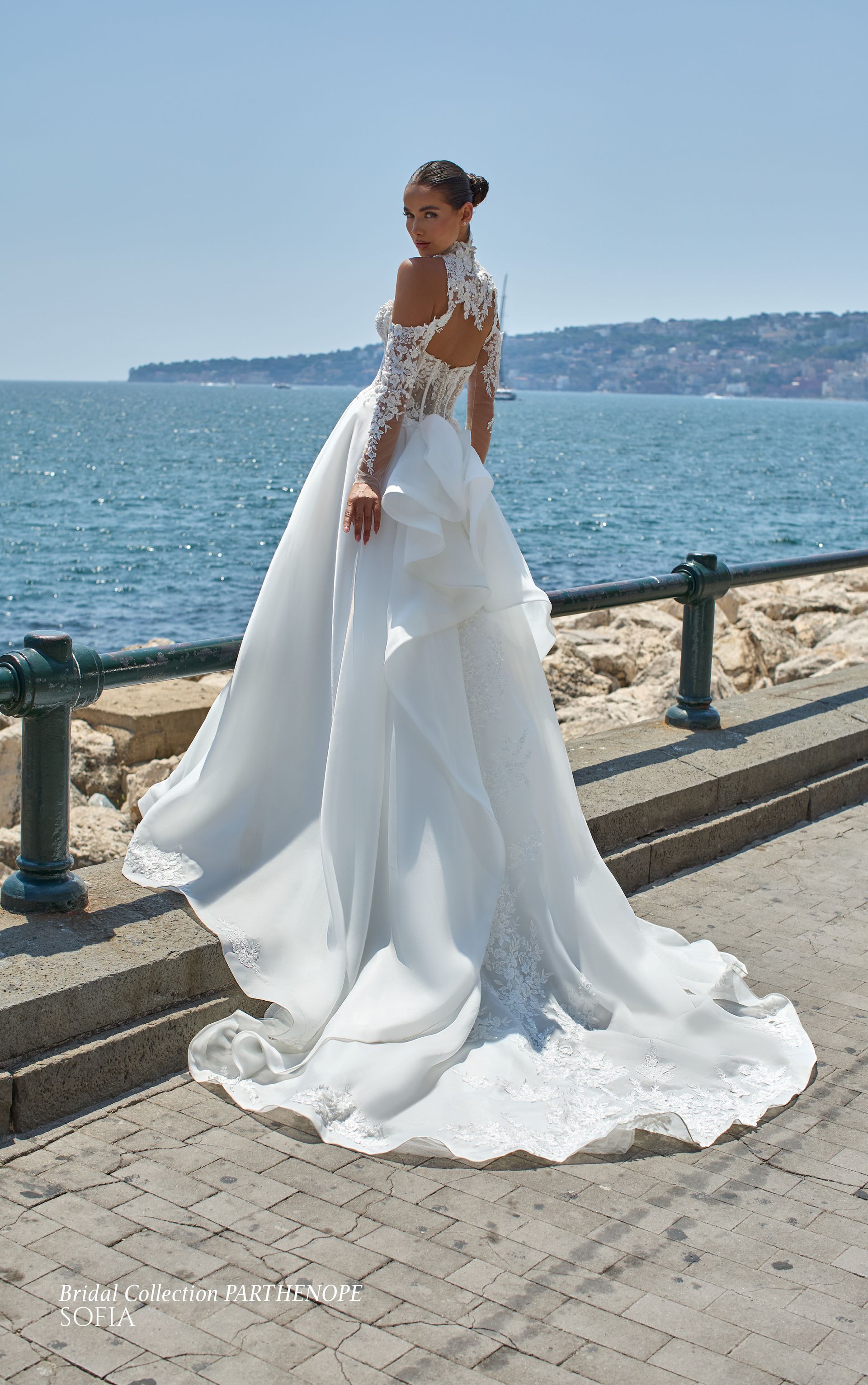 Una donna in abito da sposa bianco è in piedi in riva al mare. Maniche lunghe, schiena scoperta, strascico fluido,
