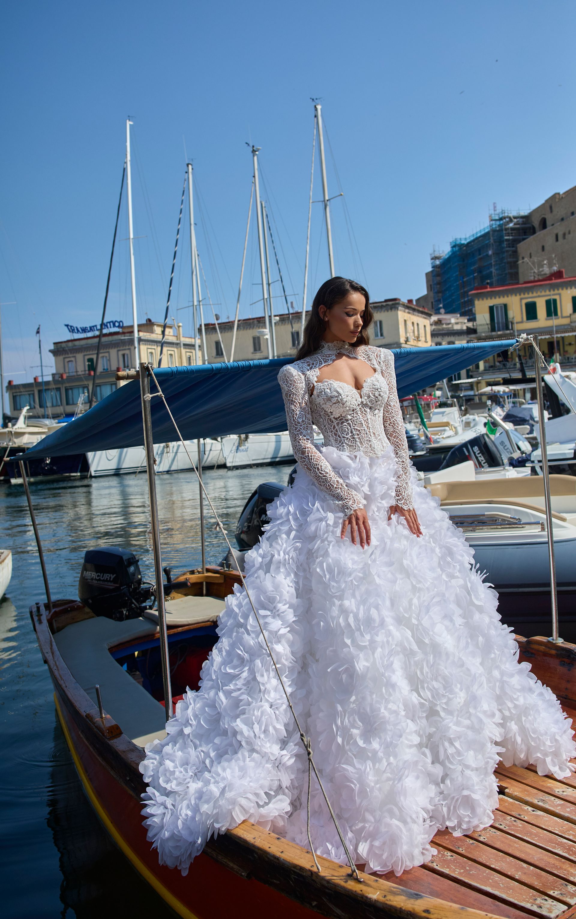 Una donna con un abito da sposa bianco con volant è in piedi su una piccola barca in un porto con barche a vela.