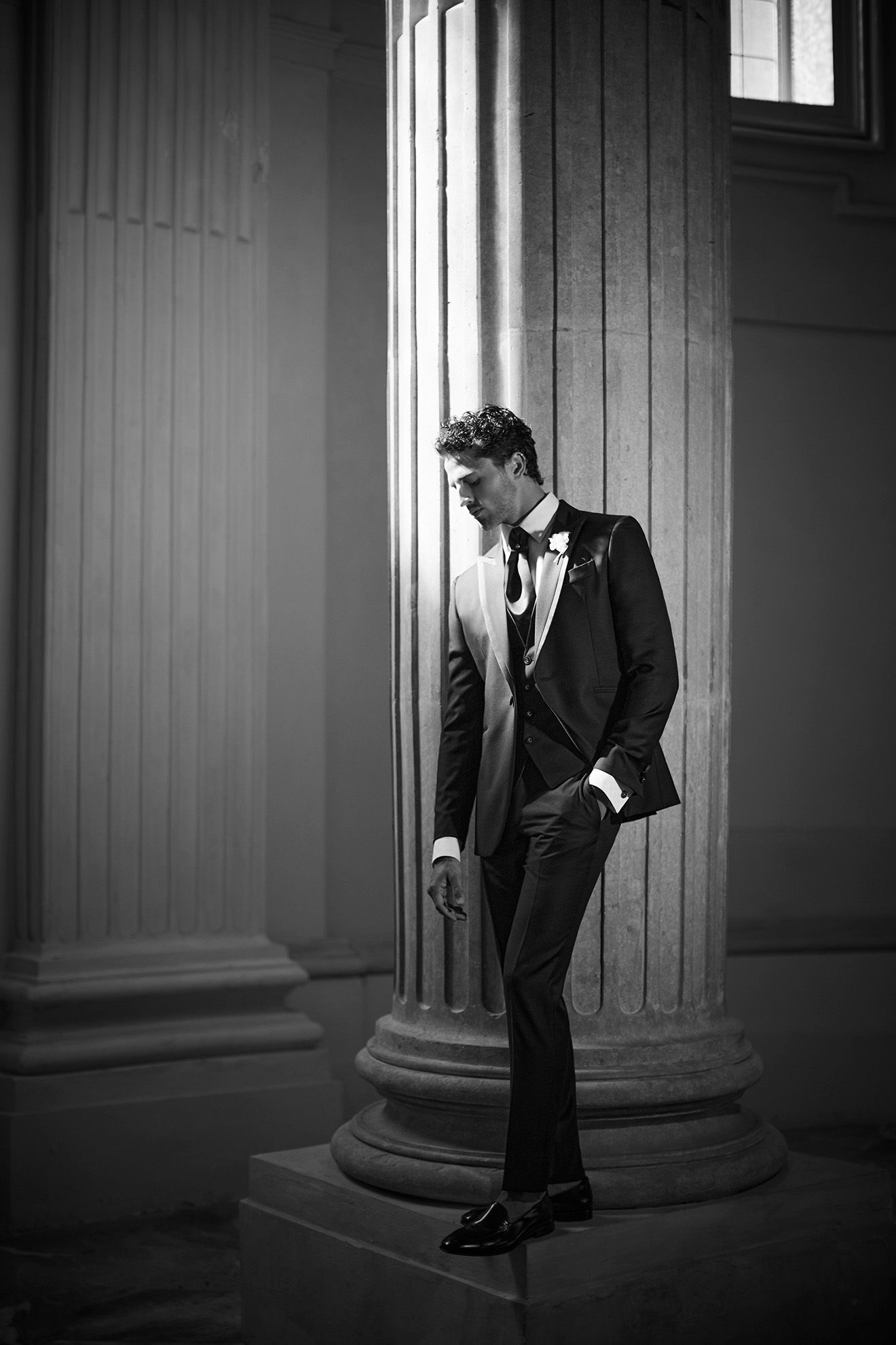 Un uomo in smoking si appoggia a un pilastro, in un contesto formale, fotografia in bianco e nero.