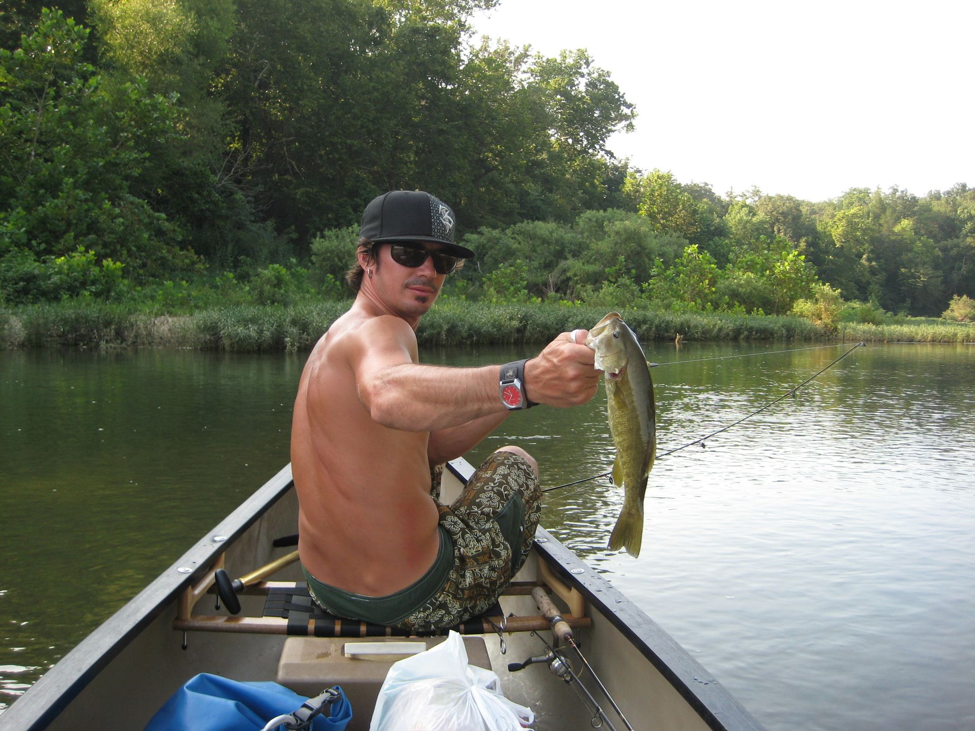 Man in a canoe holding up a fish on a calm, tree-lined river