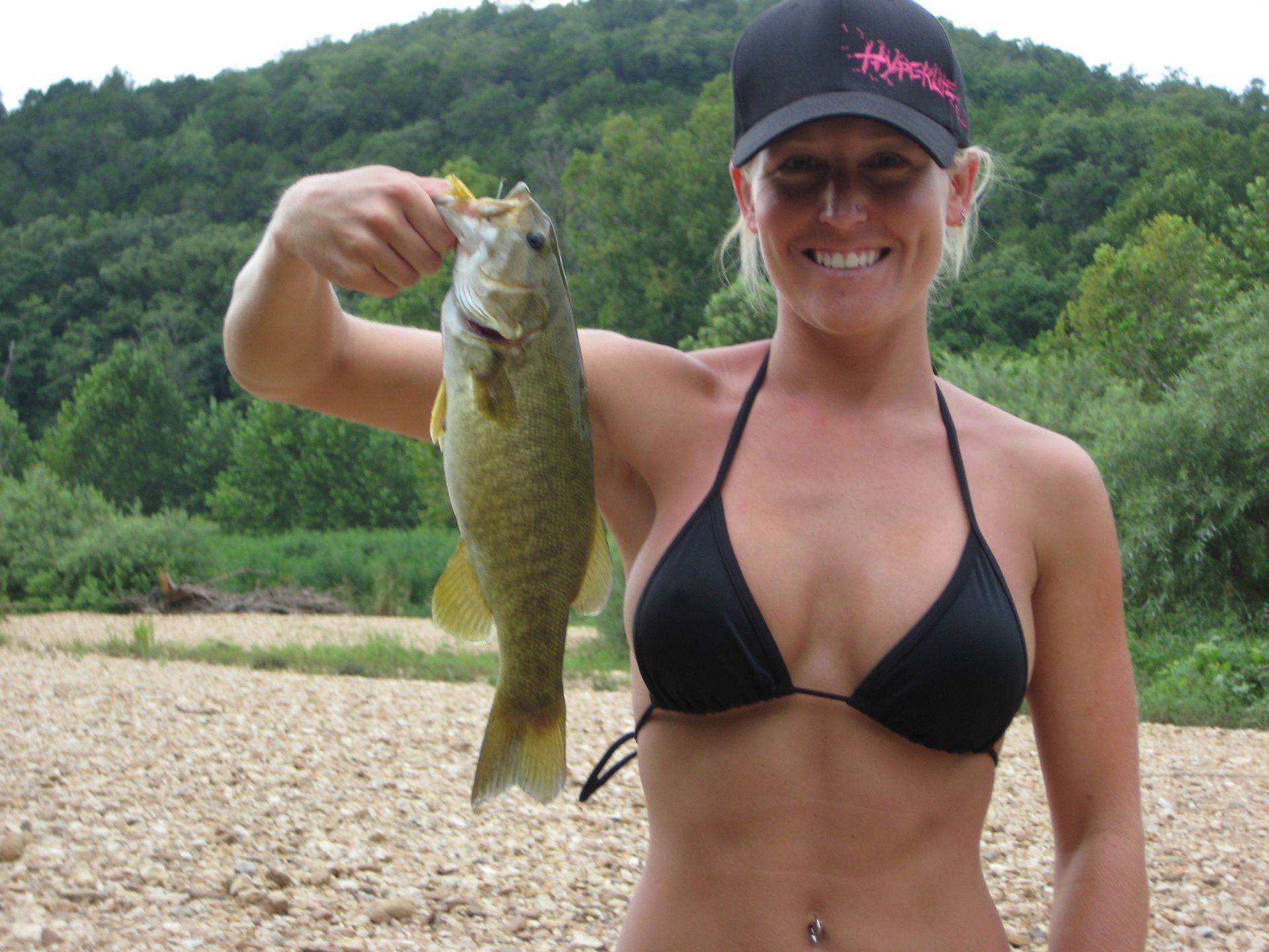 Woman holding a large fish by a riverbank, wearing a black bikini and cap, with green hills in the background